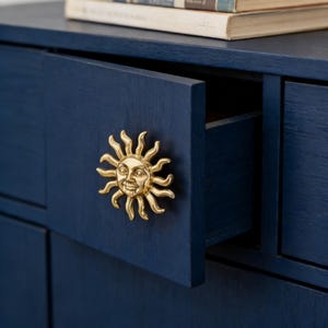 Könnte beinhalten: Ein marineblauer Schrank mit einer leicht geöffneten Schublade, die einen goldfarbenen, sonnenförmigen Griff freigibt. Der Griff hat ein lächelndes Gesicht in der Mitte. Bücher sind auf dem Schrank gestapelt.