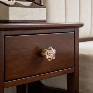 Op de afbeelding: Een donkerbruin houten nachtkastje met een stervormige, heldere glazen knop. Het nachtkastje heeft een lade en maakt deel uit van een slaapkamermeubelset. Een met stof gevoerde sieradendoos staat erop.