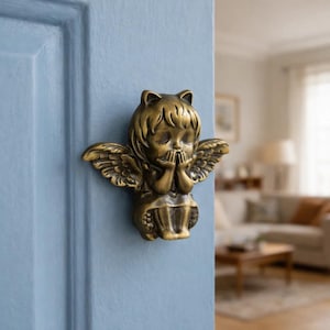 May include: A bronze-colored door knocker in the shape of a cherub with folded hands and feathered wings. The cherub is attached to a light blue door, with a blurred interior in the background.
