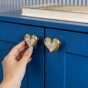Puede incluir: Dos tiradores de armario en forma de corazón con acabado dorado y centros iridiscentes. Los tiradores están montados en una puerta de armario azul. Los tiradores tienen un borde decorativo de pequeños remaches. Una mano toca uno de los tiradores.