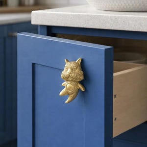 Puede incluir: Un pomo dorado con forma de gato está montado en una puerta de armario azul. El pomo es un diseño detallado en tres dimensiones. La puerta del armario está abierta, revelando un cajón de madera. El entorno de la cocina incluye una encimera blanca.