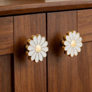 Può includere: Due pomelli per armadi a forma di margherita. Ogni pomello presenta un fiore in smalto bianco con un centro dorato e un bordo dorato. I pomelli sono montati su un mobile in legno marrone scuro.