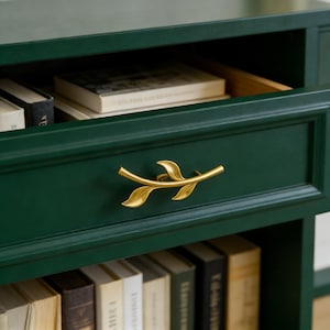 Könnte beinhalten: Ein dunkelgrüner Nachttisch mit einer Schublade mit einem goldfarbenen, blattförmigen Griff. Die Schublade ist teilweise geöffnet und gibt den Blick auf Bücher frei. Der Nachttisch hat ein Regal mit Büchern.