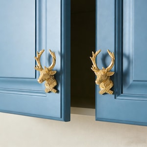 Op de afbeelding: Twee goudkleurige hertenkopkastknoppen gemonteerd op blauwe kastdeuren. De hertenkoppen hebben gedetailleerde geweien en zijn met een decoratief kruisvormig element aan de deuren bevestigd. De kastdeuren staan iets open.