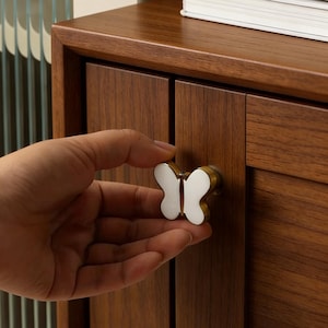 Puede incluir: Un tirador de armario con forma de mariposa con centro blanco y bordes dorados. El tirador está fijado a un armario de madera marrón oscuro. El armario tiene dos puertas y una parte superior plana. Una mano sostiene el tirador.