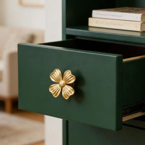Könnte beinhalten: Dunkelgrüne Schublade mit einem goldfarbenen, blütenförmigen Griff. Die Schublade ist teilweise geöffnet und gibt den Blick ins Innere frei. Der Griff ist mittig auf der Schubladenfront angebracht. Teil eines größeren Möbelstücks.