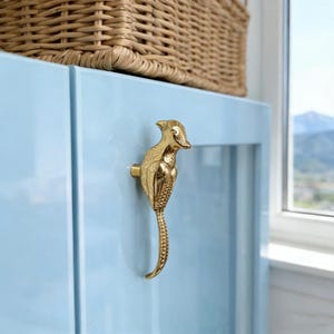 Op de afbeelding: Een goudkleurige vogelvormige kastknop is gemonteerd op een lichtblauwe kastdeur. De vogel heeft gedetailleerde veren en een lange, gebogen staart. Bovenop de kast staat een geweven mand. De achtergrond omvat een raam met uitzicht op de bergen.