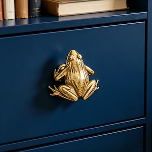 May include: A shiny gold-toned metal drawer knob shaped like a frog in a seated position. The frog figure features detailed limbs and eyes, mounted centrally on a flat, solid dark navy blue drawer front with a smooth, matte finish.