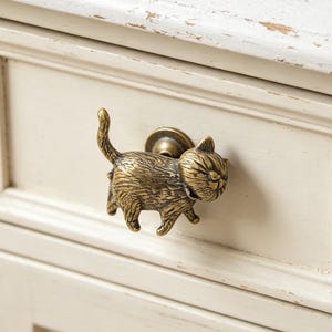 Könnte beinhalten: Ein Schubladengriff in Form einer Katze aus Messing mit detaillierter, strukturierter Oberfläche. Die Katze wird in einer Laufpose mit erhobenem Schwanz dargestellt. Der Griff ist an einer cremefarbenen Schublade befestigt und verleiht Möbeln einen skurrilen Touch.