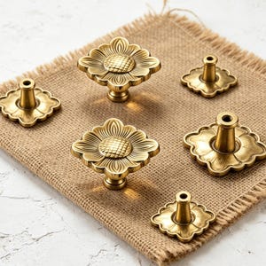 Op de afbeelding: Een set van vijf messingkleurige metalen kastknoppen, elk met een vierkant bloemmotief met een textuur in het midden en bloembladachtige randen. De knoppen zijn gerangschikt met sommige die de voorkant tonen en andere de achterste bevestigingspen.