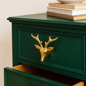 May include: A dark green dresser with a gold deer head drawer pull. The drawer is open, revealing a light brown interior. A stack of books and a white plate sit on top of the dresser.