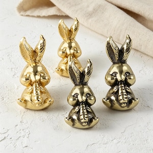 May include: Four small, polished gold-toned metal rabbit figurines, each depicted in a seated position while holding a carrot. The rabbits feature upright ears and smooth, reflective surfaces, arranged in a cluster on a textured white surface.