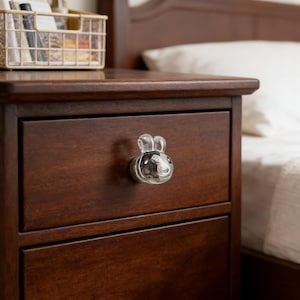 Op de afbeelding: Een close-up van een donkerbruin houten nachtkastje met twee lades. De bovenste lade heeft een heldere glazen knop in de vorm van een konijn. Een wit bed en een mand zijn op de achtergrond te zien.