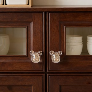 Op de afbeelding: Close-up van een donkerbruine kast met glazen deuren. Elke deur heeft een doorzichtige, beer-vormige knop. In de kast bevinden zich witte kommen en een witte container. De kast is van hout.