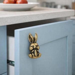 Op de afbeelding: Een messingkleurige kastknop in de vorm van een konijn is gemonteerd op een lichtblauwe kastdeur. De knop is een gedetailleerde sculptuur van een zittend konijn. De kastdeur staat iets open en onthult een witte lade binnenin.