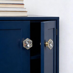 May include: A navy blue cabinet with two doors, each adorned with a clear, faceted crystal knob set in a gold-colored base. The cabinet doors are slightly ajar, revealing a dark interior. A stack of books rests on top.