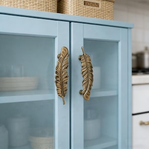 Op de afbeelding: Een lichtblauwe kast met glazen deuren en decoratieve bronzen handgrepen in de vorm van veren. De kast heeft planken met witte schalen en containers erin. Rotan manden staan bovenop de kast.