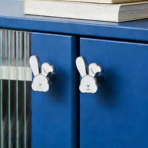 Puede incluir: Dos tiradores de puerta de armario plateados con forma de cabeza de conejo, con detalles de esmalte blanco, montados en un armario azul. Dos libros están apilados encima del armario.