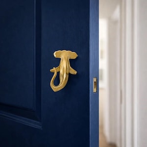 May include: A gold-colored door knocker in a unique, abstract design is mounted on a dark blue door. The knocker has a stylized, organic shape with a curved tail and a decorative top. The door also features a rectangular keyhole.