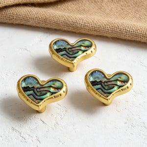 May include: Three heart-shaped cabinet knobs featuring a gold-toned metal frame with a textured edge and an inlaid iridescent abalone shell centre. Each knob has a smooth, rounded heart silhouette and a central post for mounting to furniture.