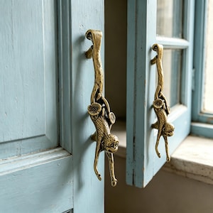 May include: Two brass monkey-shaped door handles are attached to a light blue door. The handles are detailed with a monkey hanging from a branch, with floral accents. The door is slightly ajar, revealing a glimpse of a window.