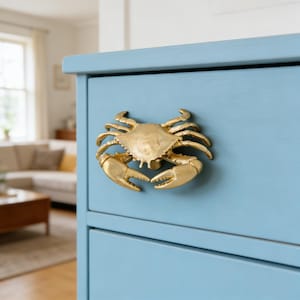 Op de afbeelding: Een goudkleurige krabvormige ladeknop is bevestigd op een lichtblauwe dressoir. Het krabontwerp is gedetailleerd, met zichtbare scharen en lichaam. De dressoir heeft een strak, modern design. De achtergrond toont een glimp van een woonkamer.