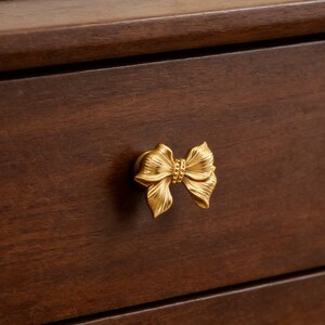 Puede incluir: Un pomo de armario dorado en forma de lazo. El pomo está fijado a un cajón de madera marrón oscuro. El lazo tiene texturas detalladas y un centro decorativo, añadiendo un toque de elegancia al mueble.