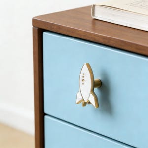 Puede incluir: Primer plano de un cajón azul claro con un tirador en forma de cohete blanco y dorado. El cajón forma parte de una cómoda de madera con la parte superior marrón. Un libro está encima.