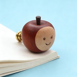 Op de afbeelding: Een houten waszegelstempel in de vorm van een appel met een lachend gezicht. De stempel is gemaakt van donkerbruin hout met een licht houten gezicht en een kleine, donkerbruine steel. De stempel ligt op een wit boek met een lichtblauwe achtergrond.