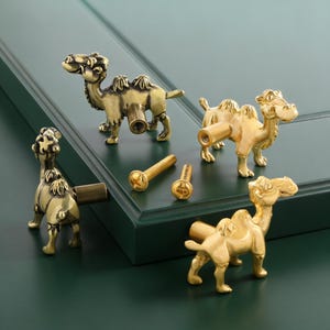 Puede incluir: Pomos de armario con forma de camello de latón y tornillos. Los pomos están detallados con jorobas y rasgos faciales, y están colocados contra una puerta de armario verde. Los tornillos son de color dorado.