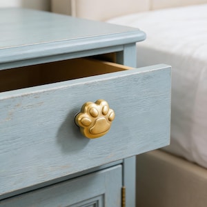 Puede incluir: Una mesita de noche azul claro con un cajón abierto, con un pomo dorado en forma de huella de pata. La mesita tiene un acabado envejecido y el cajón está vacío. El fondo incluye una cama con una colcha blanca.