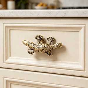 May include: A brass platypus-shaped cabinet pull is fixed to a cream-coloured drawer. The detailed, textured design of the platypus adds a unique, whimsical touch to the cabinetry. The pull is a decorative hardware element.