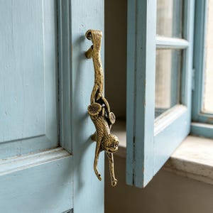 Può includere: Maniglia per porta in ottone a forma di scimmia appesa a un ramo. La maniglia è montata su una porta azzurra, con un telaio della finestra visibile sullo sfondo. La scimmia ha un volto dettagliato e si aggrappa al ramo.