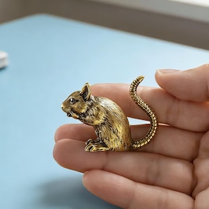 Op de afbeelding: Een gedetailleerd messing muisbeeldje met een gekrulde staart. De kleine sculptuur is goudkleurig en heeft ingewikkelde details, waaronder zichtbare oren, ogen en snorharen. De muis wordt in de hand van een persoon gehouden tegen een lichtblauwe achtergrond.