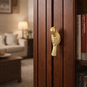 May include: A gold-coloured bird-shaped cabinet pull is mounted on a dark brown wooden cabinet door. The bird pull has detailed feather and eye features. The cabinet door is part of a larger wooden cabinet with books on a shelf.