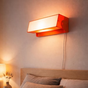 Op de afbeelding: Een oranje en witte wandlamp boven een bed. De lamp heeft een rechthoekige vorm met een naar beneden gerichte lichtbron. Het bed heeft een houten hoofdbord, kussens en een dekbed. Een lamp en vaas staan op het nachtkastje.