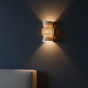 May include: A modern wall sconce with a white, hourglass-shaped design and a woven rattan center. The sconce emits warm light, casting a glow on the dark blue wall. The bedroom setting includes a bed and a small vase on a wooden nightstand.