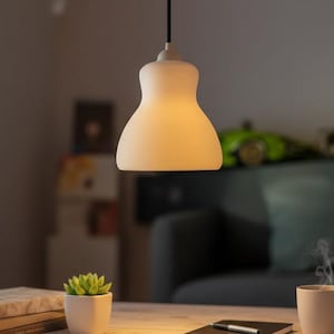 Könnte beinhalten: Eine weiße, sanduhrförmige Pendelleuchte hängt an einem schwarzen Kabel. Das warme Licht beleuchtet einen Tisch mit einer Sukkulente in einem weißen Topf, Büchern und einer Tasse Kaffee. Ein grünes Spielzeugauto steht im Hintergrund auf einem Regal.