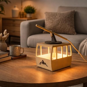 May include: A ski lift cabin-shaped table lamp with a mountain graphic. The beige lamp emits a warm, yellow glow and has a black support arm and cable. It rests on a round wooden table, with other items nearby.