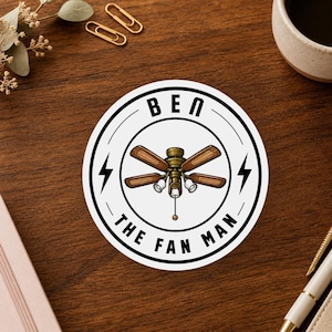 May include: A circular white sticker featuring a black border and the text BEN THE FAN MAN. In the center is an illustration of a brown ceiling fan with four blades, three light fixtures, and a pull chain, flanked by two black lightning bolts.