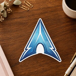 May include: A die-cut sticker featuring a stylized, triangular blue emblem with a sharp, pointed top and a central cutout. The design displays various shades of blue with a metallic, glossy finish and a distinct white border surrounding the shape.