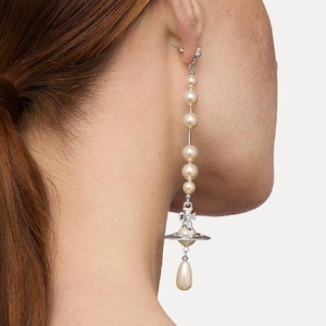 Può includere: Un orecchino lungo e pendente con perle color crema collegate da una catena d'argento. L'orecchino ha un ciondolo d'argento con una goccia di perla. L'orecchino è indossato sull'orecchio di una persona.