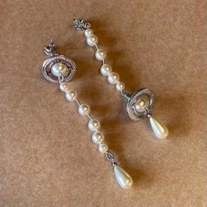 Pode incluir: Um par de brincos pendurados prateados com design de p&eacute;rolas. Cada brinco tem um pino circular com uma p&eacute;rola central, uma fileira de p&eacute;rolas e uma gota de p&eacute;rola em forma de p&ecirc;ra. Exibidos em uma superf&iacute;cie marrom.