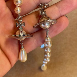 Pode incluir: Brincos prateados com detalhes de p&eacute;rolas. Um brinco tem um design de cruz com pedras preciosas coloridas, uma p&eacute;rola e uma p&eacute;rola em forma de gota. O outro brinco tem um design semelhante a um planeta com uma p&eacute;rola e uma fileira de p&eacute;rolas.