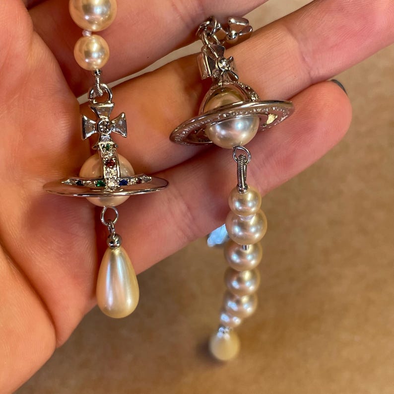 Pode incluir: Brincos prateados com detalhes de p&eacute;rolas. Um brinco tem um design de cruz com pedras preciosas coloridas, uma p&eacute;rola e uma p&eacute;rola em forma de gota. O outro brinco tem um design semelhante a um planeta com uma p&eacute;rola e uma fileira de p&eacute;rolas.
