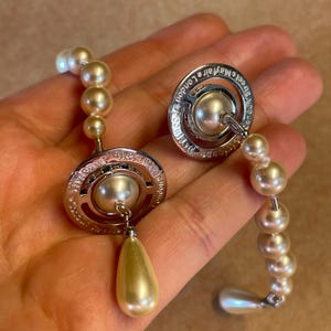 Pode incluir: Brincos prateados com uma grande p&eacute;rola central e um design circular gravado. Cada brinco tem uma gota de p&eacute;rola em forma de l&aacute;grima e &eacute; conectado por uma corrente de p&eacute;rolas menores. O texto 'Vivienne Westwood Mayfair London' &eacute; vis&iacute;vel.
