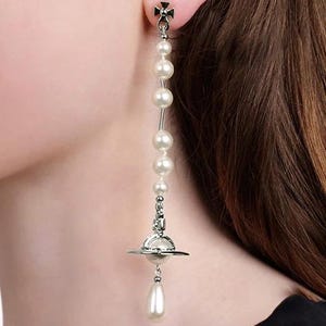 Pode incluir: Um brinco longo e pendurado com um design de planeta prateado e uma gota de p&eacute;rola. O brinco apresenta uma s&eacute;rie de p&eacute;rolas brancas e redondas enfiadas ao longo de uma haste prateada. A parte superior do brinco tem um design de cruz.