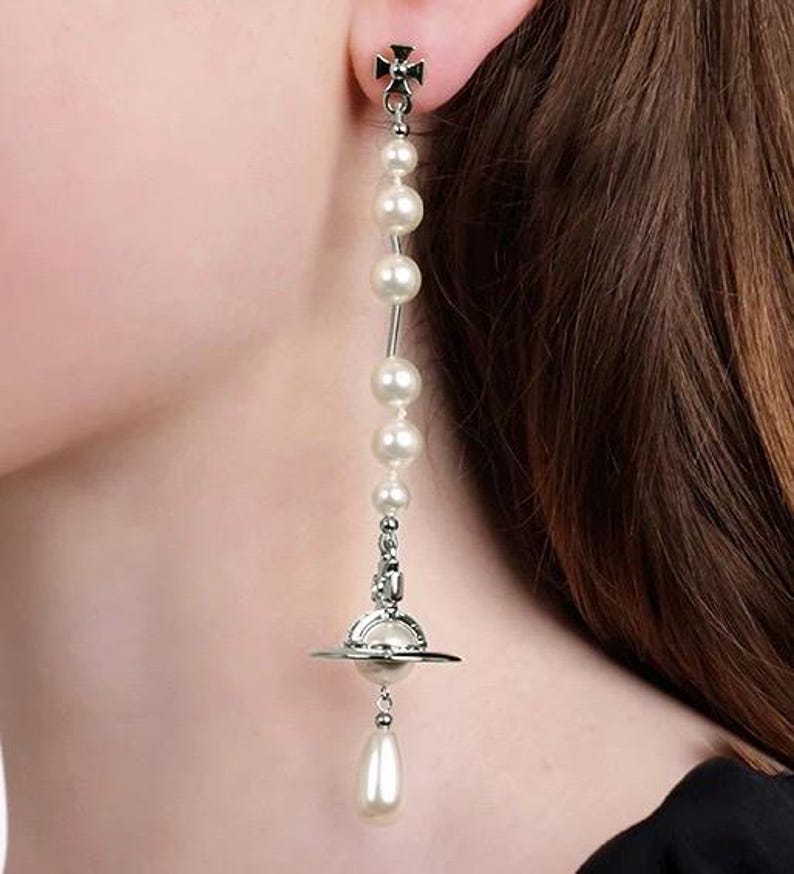 Pode incluir: Um brinco longo e pendurado com um design de planeta prateado e uma gota de p&eacute;rola. O brinco apresenta uma s&eacute;rie de p&eacute;rolas brancas e redondas enfiadas ao longo de uma haste prateada. A parte superior do brinco tem um design de cruz.
