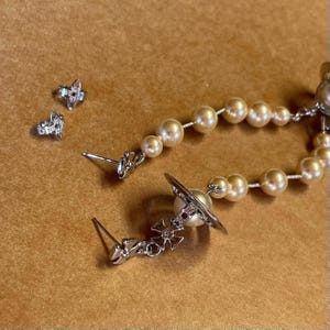 Pode incluir: Um conjunto de brincos prateados e um colar com um fio de p&eacute;rolas. Os brincos incluem brincos de press&atilde;o e brincos pendurados com um design de planeta. O colar apresenta um fio de p&eacute;rolas de cor creme.