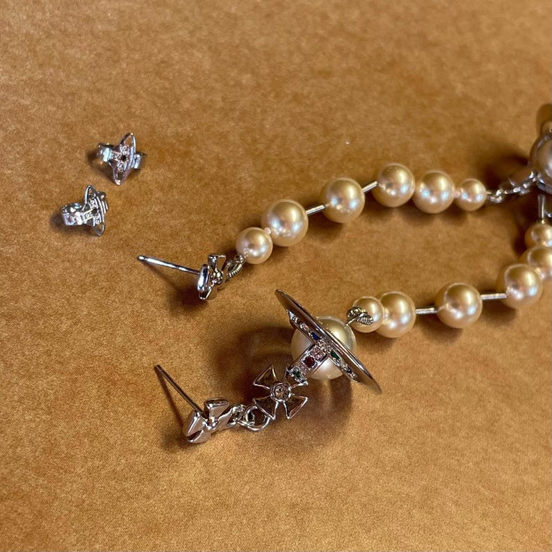 Pode incluir: Um conjunto de brincos prateados e um colar com um fio de p&eacute;rolas. Os brincos incluem brincos de press&atilde;o e brincos pendurados com um design de planeta. O colar apresenta um fio de p&eacute;rolas de cor creme.